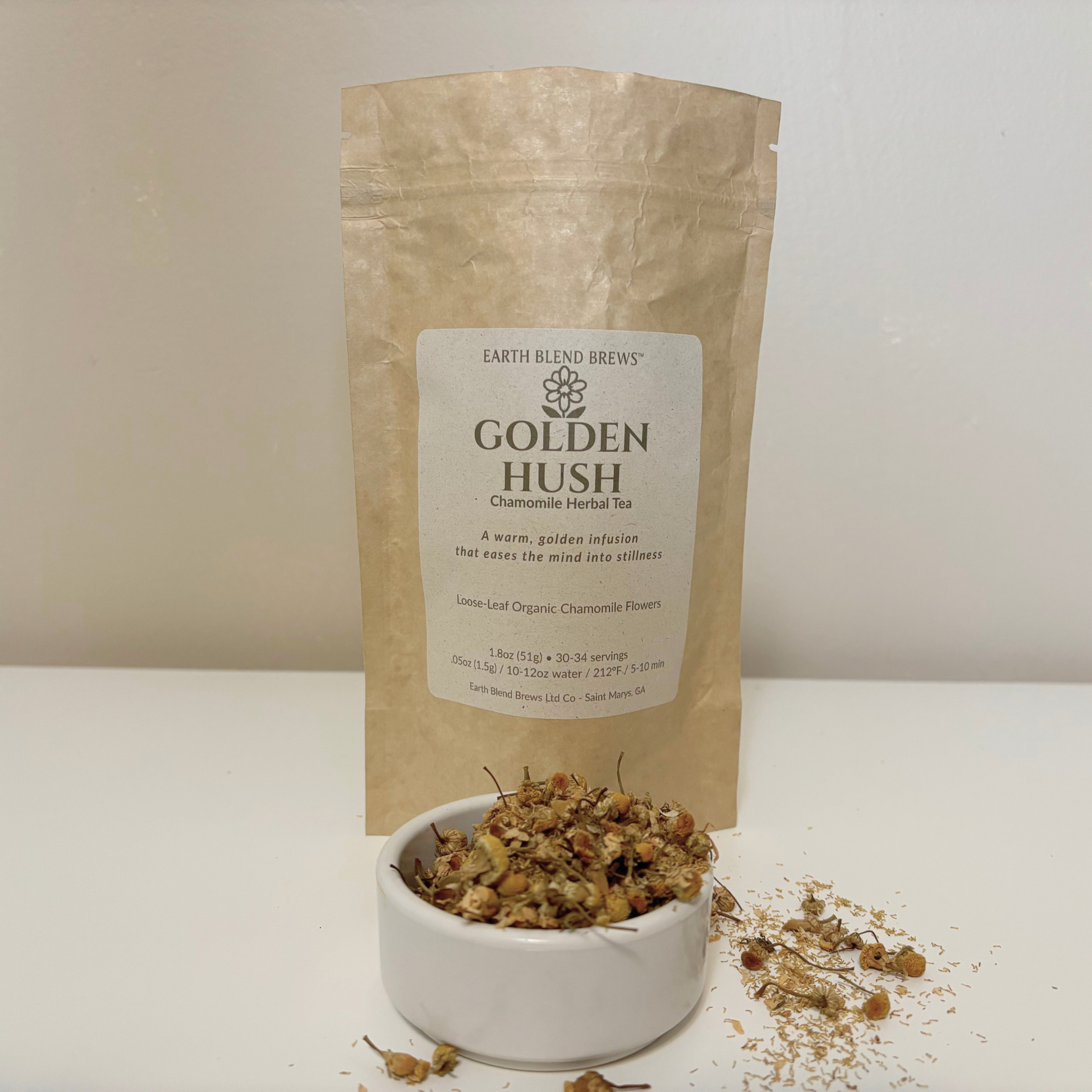 Bag of Golden Hush Chamomile Herbal Tea from Earth Blend Brews with a small bowl of tea leaves on a white surface.
