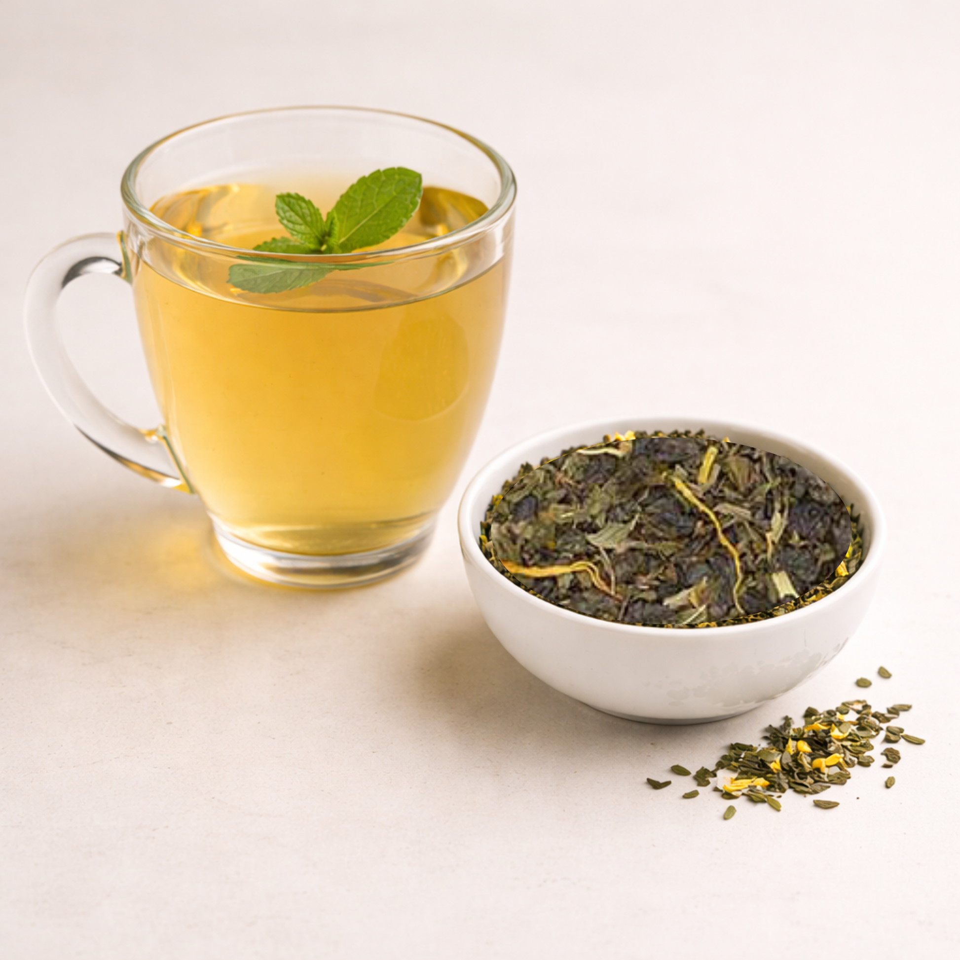 Glass of green tea with a mint leaf, bowl of green tea leaves, and scattered tea leaves on a light surface.