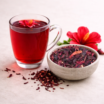 Glass of red hibiscus tea with a bowl of dried hibiscus flowers and a red hibiscus flower on a light background.